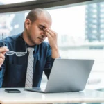 A person sitting in a chair with a worried expression, holding their head and looking ill, surrounded by a blurred office environment in the background. Representing Sick Building Syndrome.