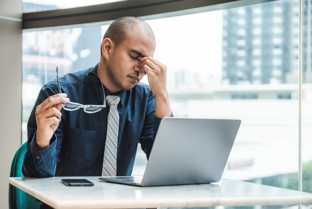 A person sitting in a chair with a worried expression, holding their head and looking ill, surrounded by a blurred office environment in the background. Representing Sick Building Syndrome.